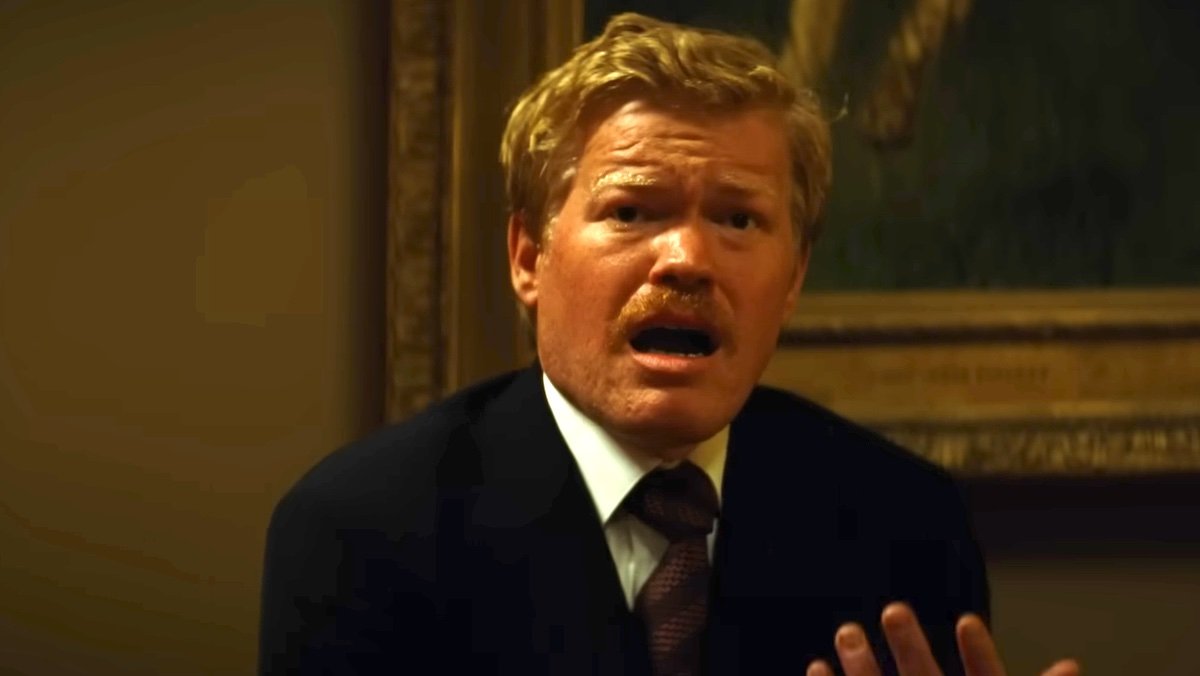 A mustachioed JEsse Plemons in a suit on the ground yelling in pain while holding his hand in Kinds of Kindness