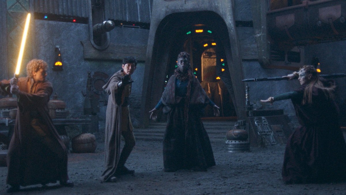 A Jedi draws his yellow lightsaber to fight a witch with a staff as anaother Jedi and witch stand between them on The Acolyte